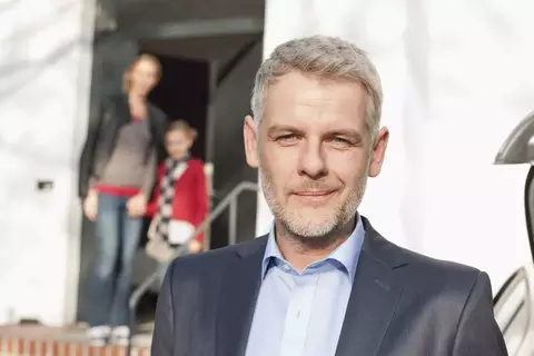 Afbeelding van een man in kostuum, met op de achtergrond een vrouw en kind in de deuropening