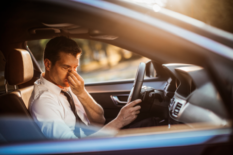 Afbeelding van een man aan het stuur van een auto, terwijl die 1 hand voor zijn gezicht houdt