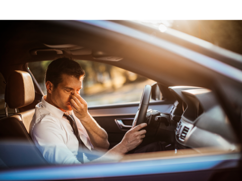 Afbeelding van een man aan het stuur van een auto, terwijl die 1 hand voor zijn gezicht houdt